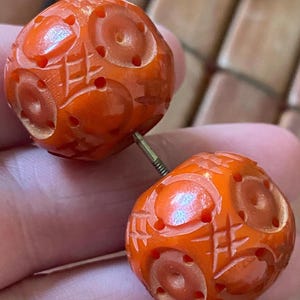 May include: A pair of orange, carved beads connected by a metal pin. Each bead is spherical with a detailed pattern of circles and lines. The beads have a glossy finish and are held between fingers.