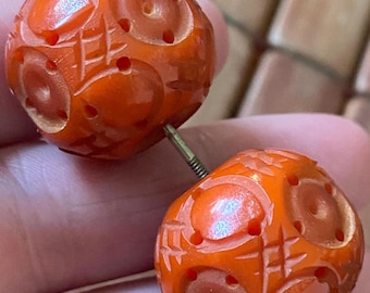 May include: A pair of orange, carved beads connected by a metal pin. Each bead is spherical with a detailed pattern of circles and lines. The beads have a glossy finish and are held between fingers.