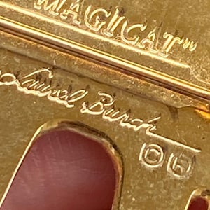 May include: Gold-toned metal brooch with the inscription "MAGICAT" and "Laurel Burch". The brooch has a cat-like silhouette with cut-out details. The design includes copyright symbols.