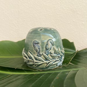 May include: A teal ceramic flower frog with three holes on top. The sides feature two seahorse figures and leafy designs in a lighter shade of teal. The frog is sitting on a large green leaf.