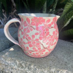 May include: A ceramic jug with a handle and spout, adorned with pink cherry blossom patterns on a cream backdrop. The jug is placed on a grey stone surface.