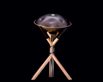 May include: A dark gray steel handpan with a wooden stand. The handpan is resting on a tripod stand made of light brown wood.