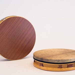 May include: Two wooden frame drums, one with a brown wooden surface and the other with a light brown leather surface. The leather drum has a black rim and a decorative wooden base.