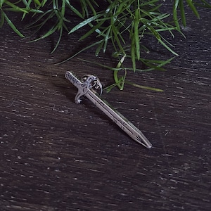 May include: A silver sword pin with a detailed design. The pin is on a dark wood surface.