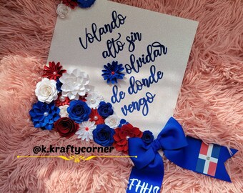 May include: A white graduation cap decorated with blue and red paper flowers, a blue bow, and a Dominican Republic flag. The cap has the text "Volando alto sin olvidar de donde vengo" and "@k.kraftycorner" written on it. The cap also has the text "FHHS 21" written on a blue ribbon.