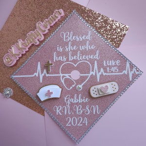 May include: Pink graduation cap with white and gold accents. The cap has a stethoscope, a bandage, a cross, and a nurse's hat. The text on the cap reads "Blessed is she who has believed Luke 1:45" and "Gabbie RN, BSN 2024".
