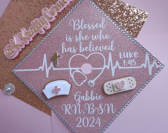 May include: Pink graduation cap with white and gold accents. The cap has a stethoscope, a bandage, a cross, and a nurse's hat. The text on the cap reads "Blessed is she who has believed Luke 1:45" and "Gabbie RN, BSN 2024".