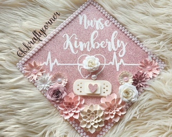 May include: Pink graduation cap decorated with white pearls, white and pink paper flowers, a white bandage with a red heart, and a white heartbeat graphic with the text "Nurse Kimberly".