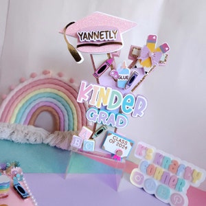 May include: A pastel rainbow cake topper with the words "Kinder Grad" and "K's Krafty Corner" in rainbow colors. The topper also includes a pink graduation cap with the name "Yannetley" on it, a book with the words "Class of 2024", and other school-themed decorations.