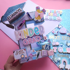 May include: A blue graduation cap decorated with a pink and white bow, pencils, crayons, and the words "ELIANNYS" and "KINDER GRAD". The cap also includes a small sign that says "CLASS OF 2024".