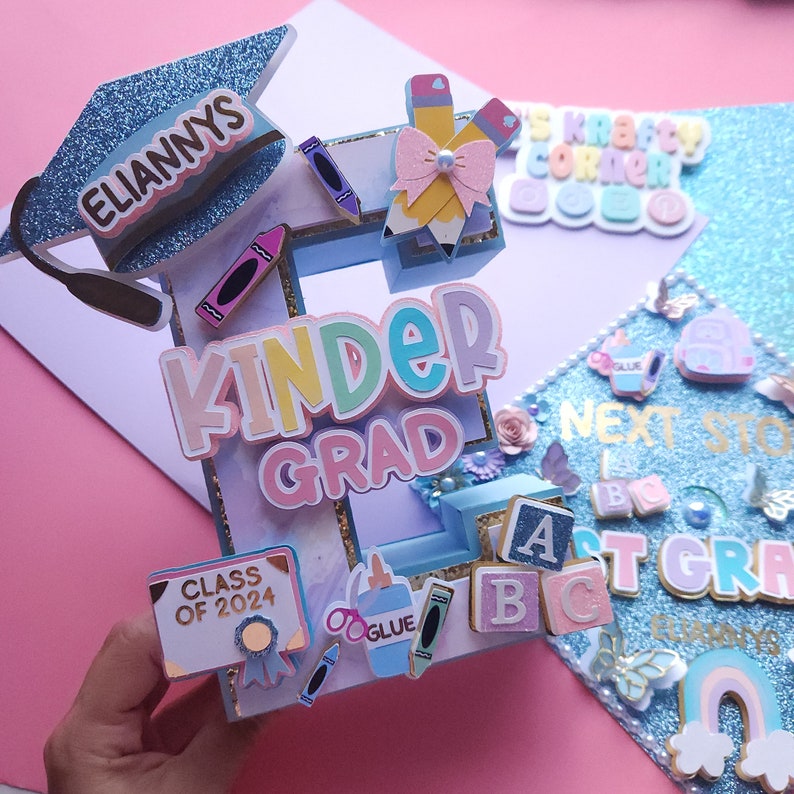 May include: A blue graduation cap decorated with a pink and white bow, pencils, crayons, and the words "ELIANNYS" and "KINDER GRAD". The cap also includes a small sign that says "CLASS OF 2024".