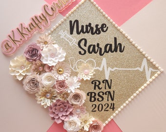 May include: A gold glitter graduation cap decorated with paper flowers in shades of pink, cream, and gold. The cap features the text "Nurse Sarah RN BSN 2024" and a syringe graphic.