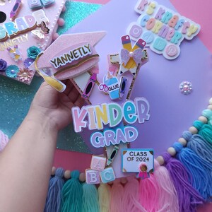 May include: A pink graduation cap with the word "Yannetly" on it, a rainbow "Kinder Grad" banner, and a blue sign that says "Class of 2024". The cap is decorated with colorful pencils, crayons, and a glue bottle. There are also colorful tassels and wooden blocks with letters on them.