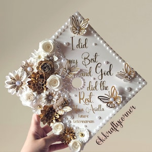 Könnte beinhalten: Eine weiße Abschluss-Kappe, verziert mit weißen und goldenen Papierblumen, Perlen und goldenen Schmetterlingen. Die Kappe trägt den Text "I did my best and God did the rest. Alexa Ariella Future Veterinarian @k.kraftycorner"