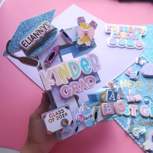 May include: A blue graduation cap with the name "Eliannys" on it, decorated with a blue and white letter "E" with the words "Kinder Grad" on it. The letter is decorated with colorful crayons, a book that says "Class of 2024", and other school-themed decorations.