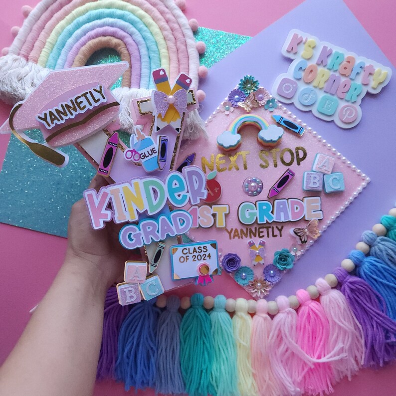 May include: A pink graduation cap decorated with colorful embellishments, including a rainbow, a graduation cap, and the words "Kinder Gradist 1st Grade". The cap is surrounded by colorful tassels and a rainbow-colored yarn garland.