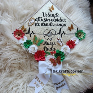 Op de afbeelding: Een gouden afstudeerhoed versierd met bloemen, vlinders en een stethoscoop. De hoed heeft de tekst "Volando alto sin olvidar de donde vengo" en "Nurse Linda" erop. De hoed heeft ook de tekst "BSN, RN 2022" en "Para mi familia" op een wit lint.