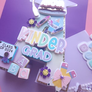 May include: A purple, pink, and blue letter A with the word "Kinder Grad" in rainbow colors. The letter A is decorated with a graduation cap, crayons, a glue bottle, and ABC blocks.