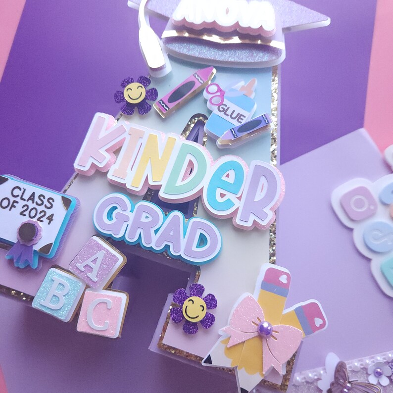 May include: A purple, pink, and blue letter A with the word "Kinder Grad" in rainbow colors. The letter A is decorated with a graduation cap, crayons, a glue bottle, and ABC blocks.