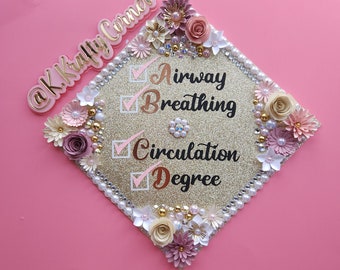 May include: A gold glitter graduation cap decorated with paper flowers, pearls, and the text "Airway Breathing Circulation Degree." The cap has a border of pearls and is embellished with pink, white, and gold flowers. The @K Krafty Corner logo is visible.