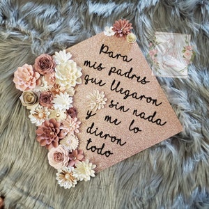 May include: A rose gold graduation cap decorated with paper flowers in shades of pink and white. The cap has a message written in black ink that reads "Para mis padres que llegaron sin nada y me dieron todo".