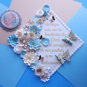 May include: A white graduation cap decorated with blue and white paper flowers, pearls, and gold butterflies. The cap has a quote in Spanish that reads: "Un logro más con la bendición de Dios, el sacrificio de mis padres y mi dedicación."