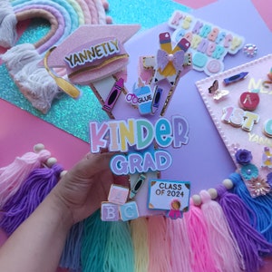 May include: A pink graduation cap with the name "YANNETLY" on it, decorated with colorful school supplies and the words "KINDER GRAD" in rainbow colors. The cap is surrounded by colorful yarn tassels and a sign that says "CLASS OF 2024".