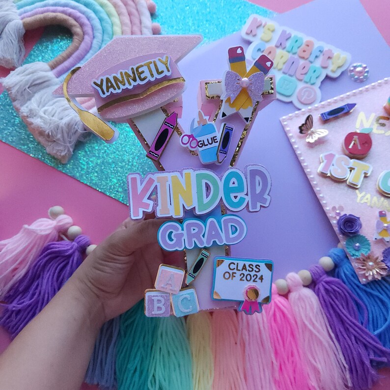 May include: A pink graduation cap with the name "YANNETLY" on it, decorated with colorful school supplies and the words "KINDER GRAD" in rainbow colors. The cap is surrounded by colorful yarn tassels and a sign that says "CLASS OF 2024".