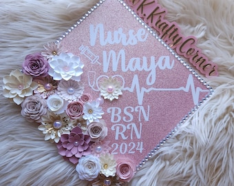 May include: Pink graduation cap decorated with white and pink paper flowers, a stethoscope, a syringe, and a heartbeat graphic. The text "Nurse Maya BSN RN 2024" is written on the cap. A pink tag with the text "@KraftyCorner" is attached to the cap.