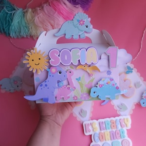 Puede incluir: Una caja de regalo blanca decorada con recortes de dinosaurios, el nombre "Sofia" y el número "1". La caja presenta un sol sonriente, flores y mariposas. La caja está decorada con colores pastel y sobre un fondo rosa.