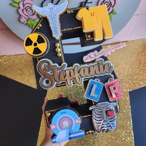 May include: Black and gold decorated number two cake topper with various medical symbols, including a stethoscope, a radiation symbol, an X-ray, a ribcage, and a scrubs top. The name "Stefanie" is written in gold lettering and the letters "LR" are on blue and red tags.