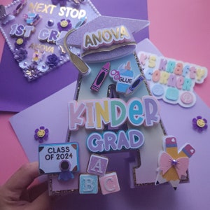 May include: A purple letter A decorated with glitter, a graduation cap, and school supplies. The letter says "Kinder Grad" and "Class of 2024".
