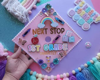 May include: Pink graduation cap decorated with colorful felt letters spelling "Next Stop 1st Grade" and other school-themed embellishments, including a rainbow, clouds, butterflies, and a red apple.