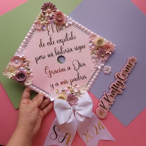 Op de afbeelding: Een roze afstudeerhoed versierd met bloemen, parels en strass. De hoed heeft een gouden inscriptie met de tekst "El fin de este capitulo pero mi historia sigue Gracias a Dios y a mis padres" en "SA 2024". De hoed is ook versierd met een witte strik en een gouden bord met de tekst "@K.Krafty Corner".