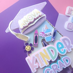 May include: A purple glitter graduation cap with the name "ANOVA" in gold letters. The cap is decorated with a gold tassel and is sitting on top of a blue and white background with the words "KINDERGARD" in rainbow letters. There are also several colorful school supplies, including a crayon, glue bottle, and a pair of glasses.