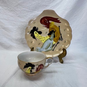 May include: A vintage teacup and saucer set with a rooster design. The cup and saucer are both white with a cream-colored background. The rooster design is in black, yellow, red, and blue.