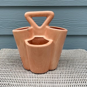 May include: A peach-colored ceramic utensil holder with a handle. The holder has three compartments and a smaller pocket in the front. The holder is sitting on a white wicker surface.