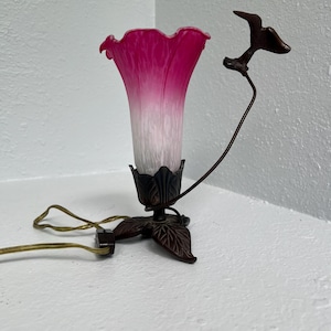 May include: A decorative table lamp with a pink and white glass shade shaped like a flower. The lamp has a bronze-colored base with leaf details and a bird accent. The lamp is plugged into an electrical outlet.