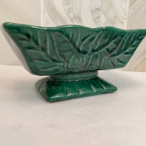 May include: A rectangular ceramic planter with a deep forest green glaze and a glossy finish. The exterior features a molded leaf pattern with raised veins. The vessel is supported by a flared, stepped pedestal base with vertical ridges.