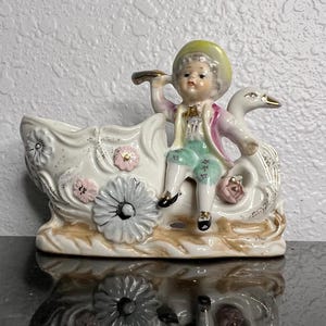 May include: A porcelain figurine of a boy in a pink and green outfit riding a swan with floral details. The swan is part of a planter with a white and gold floral design.
