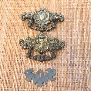 May include: Two ornate, antique brass drawer pulls with a matching keyhole escutcheon. The pulls feature intricate scrollwork and a central shield design. The hardware is set against a woven, light brown background.