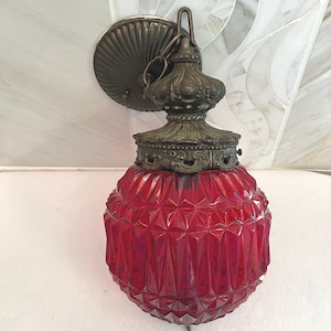 May include: A wall-mounted light fixture featuring a deep red, faceted glass globe with a diamond-cut pattern. The lamp is topped with an ornate, dark bronze-toned metal cap and a matching circular wall plate, connected by a short metal chain link.