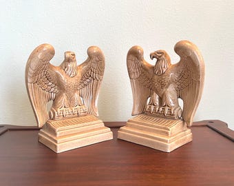 May include: A pair of tan-colored eagle bookends. Each bookend features a detailed sculpture of an eagle with outstretched wings, perched atop a rectangular base. The bookends are made of a ceramic material and are designed to hold books.