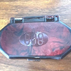 May include: A burgundy and black marbled compact case with a black frame. The case has a rectangular shape with rounded edges and a clasp closure. The number "969" is embossed on the lid.