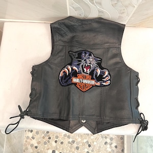 May include: A black leather vest featuring side lacing and a large embroidered patch on the back. The patch depicts a snarling black panther with white claws above the orange and white text reading MOTOR HARLEY-DAVIDSON CYCLES.
