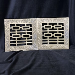 May include: Two square brass panels featuring a central geometric openwork pattern composed of rectangular bars. Each panel is framed by a decorative border embossed with a repeating leaf motif. The metal surfaces show a muted, aged finish.