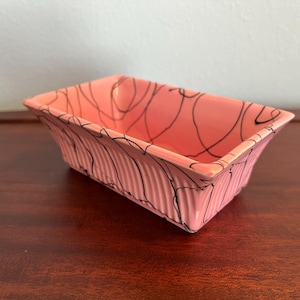 May include: A rectangular, pink ceramic dish with a ribbed exterior and black abstract lines. The dish is approximately 8 inches long, 4 inches wide, and 3 inches tall. The interior is smooth, while the exterior has a textured, ridged design.