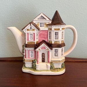 May include: A ceramic teapot shaped like a multi-story house with pink siding, white trim, and a brown roof. The structure features a central front door, multiple windows, a pointed turret, a white spout on the left, and a curved handle on the right.