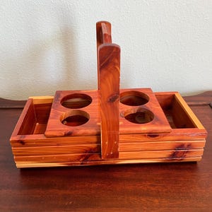 May include: A wooden beverage carrier with a handle, featuring four circular openings for holding glasses or bottles. The carrier is made of reddish-brown wood and has two side compartments.