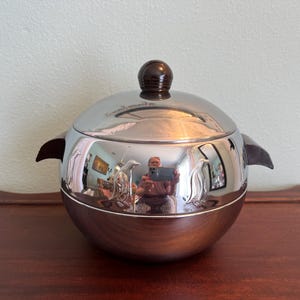 May include: A stainless steel ice bucket with a dark brown wooden base and handle. The lid is topped with a dark brown wooden knob. The bucket reflects the surrounding environment.